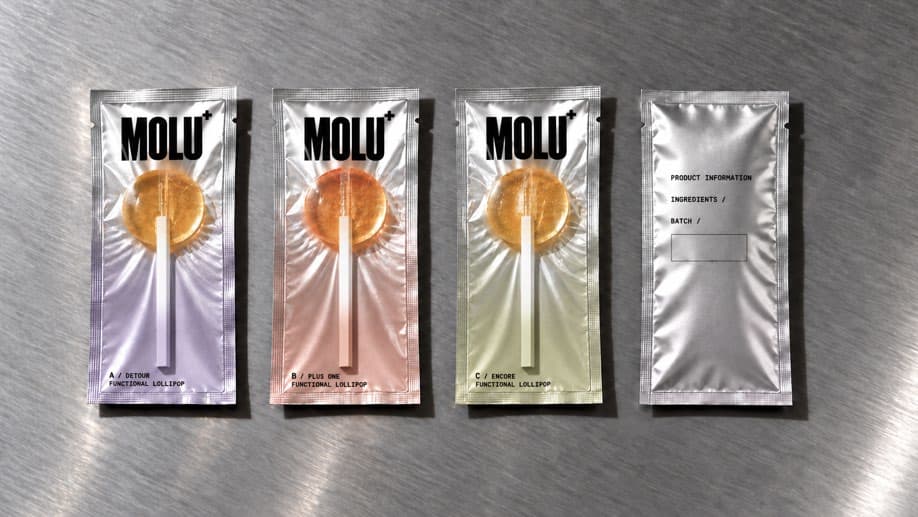Die drei MOLU+ Verpackungen A, B und C in Flieder, Peach und Olive neben der silbernen Rückseite auf gebürstetem Metall